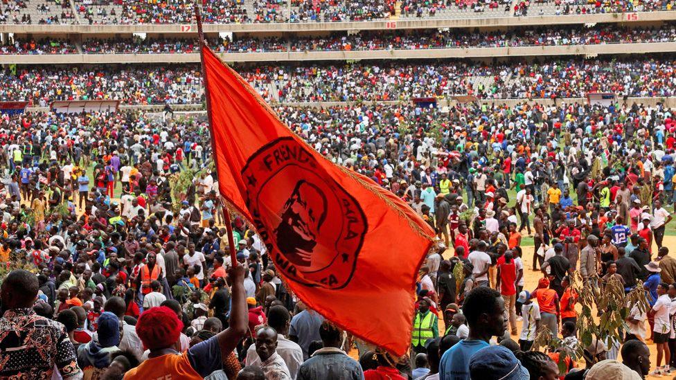 Thousands of mourners inside a stadium in Nairobi, one carrying  a massive orange flag of Raila Odinga.
