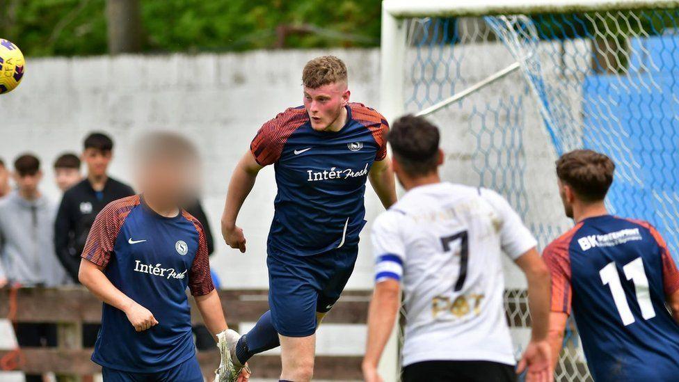 John McKenna heads a ball during a football match. he is a young man with short blonde hair wearing a navy football kit with orange patterned sleeves.