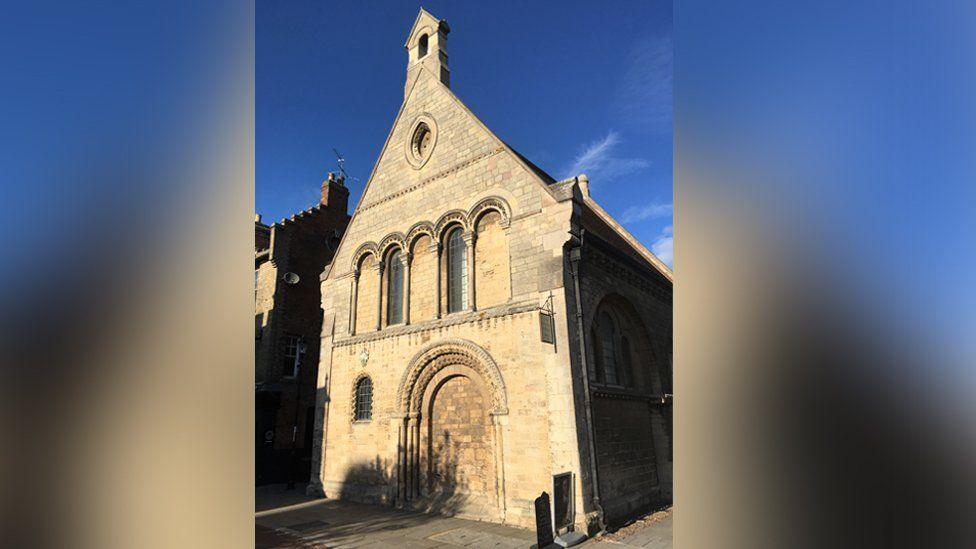 The Cromwell Museum in Huntingdon. It is a stone built 12th Century building with an arched doorway on the ground floor. The door has been bricked up. Above it is a row of arched windows, only two of which have glass. Above that is a peaked gable. The sky above is blue.