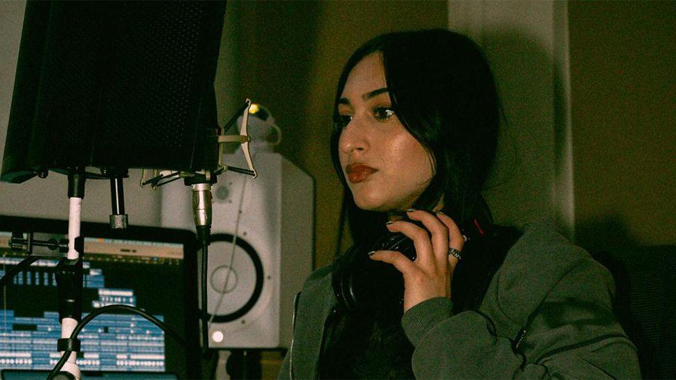 Khushi Kaur, a female singer in a music studio, looking into a microphone while holding a black pair of headphones around her neck. She is wearing a green jacket, with a computer screen and white speakers in the background.