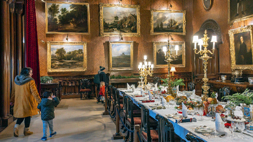 Visitors in winter coats and hats walk through the castle dining room complete with a dining table set for a feast with portraits and landscape paintings hanging from its walls