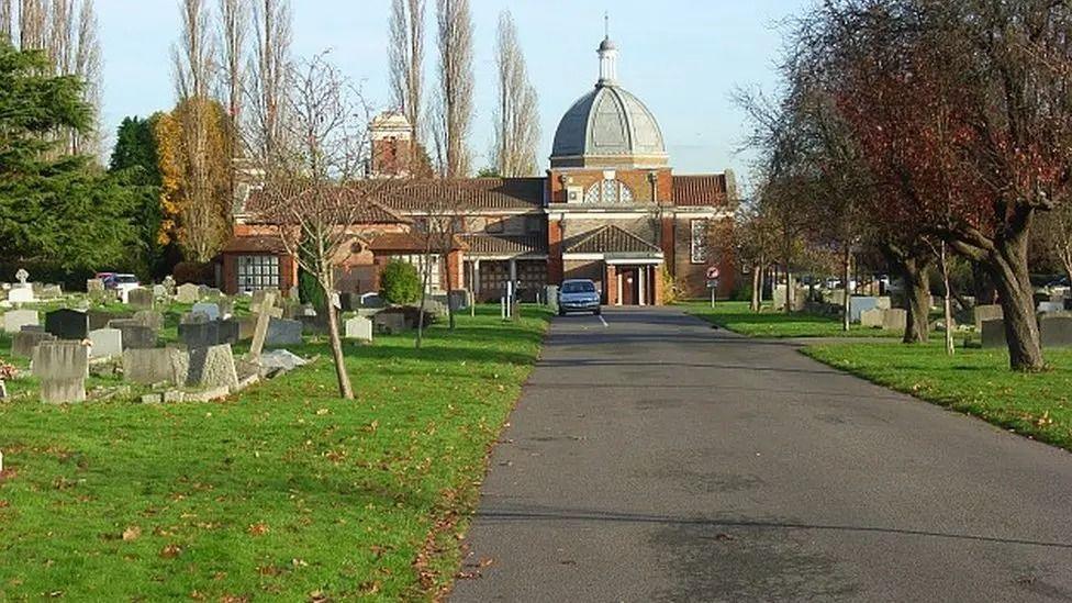 A tree-lined road through a graveyard with a red-bricked domed building in the background