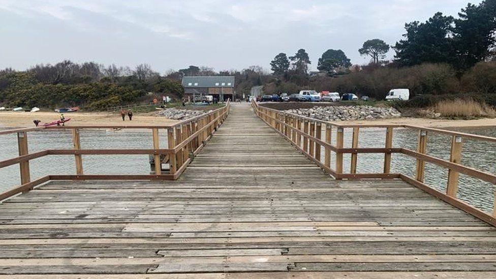 Wooden pier with planks across its width and wooden railing stretching down the passageway leading to a beach with sand and large rocks on its slope and a car park beyond.