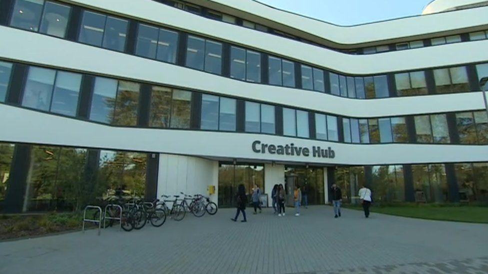 Four-storey white and glass teaching building with windows across the whole width of each storey.  The words "creative hub" are above the entrance, and there are some bicycles parked outside. A number of students are entering the building.