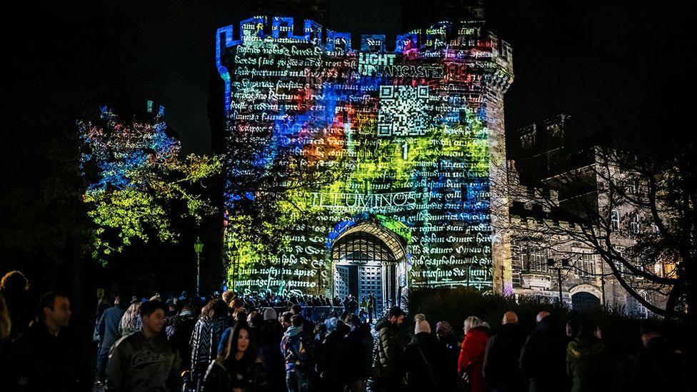 The outside of Lancaster Castle at night lit up with mediaeval writing in white on blue, orange and yellow lights. There are lots of people milling about in front 