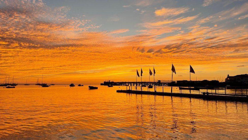 Orange and gold sky reflecting in the sea with a pontoon, flags and boats in dark sillouhette 