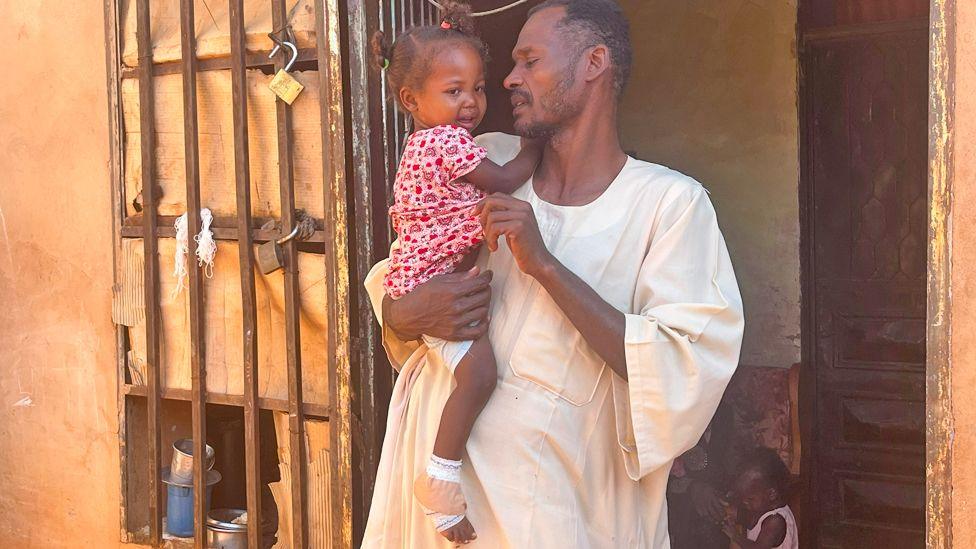 Omnia, a little girl in a red flower print dress, is held by her uncle Abazar Abdel Habib in a white dishdasha. Her foot is bandaged.