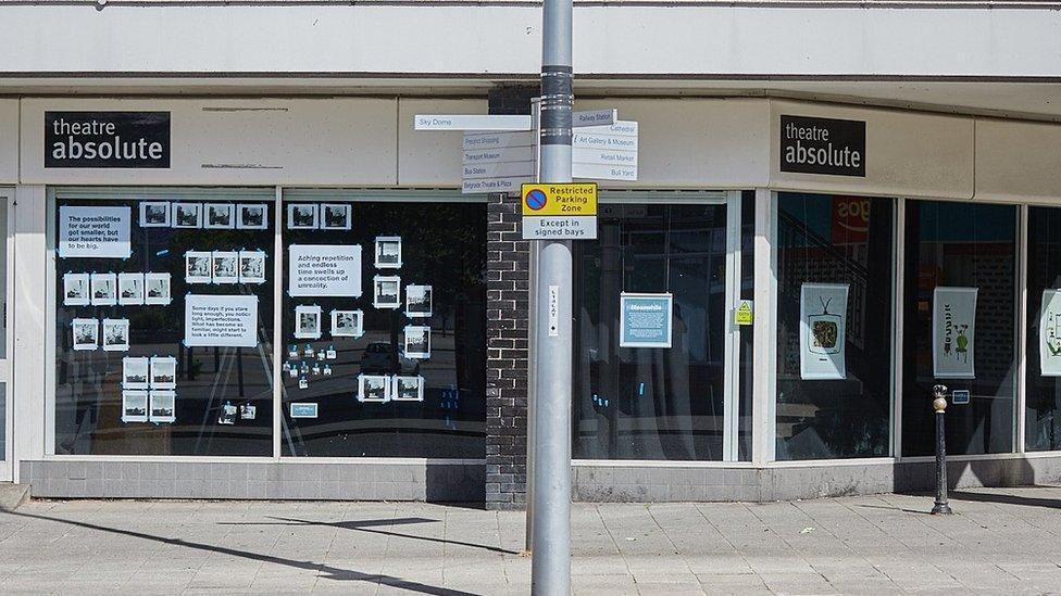 The outside of a shop front, with photos and writing stuck in the window. The signage says Theatre Absolute in white text on a black background.