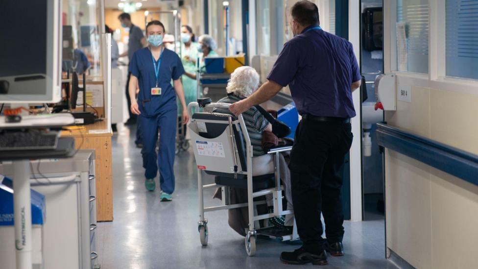 Woman walking in scrubs and man pushing woman in wheeelchair in hospital corridor.