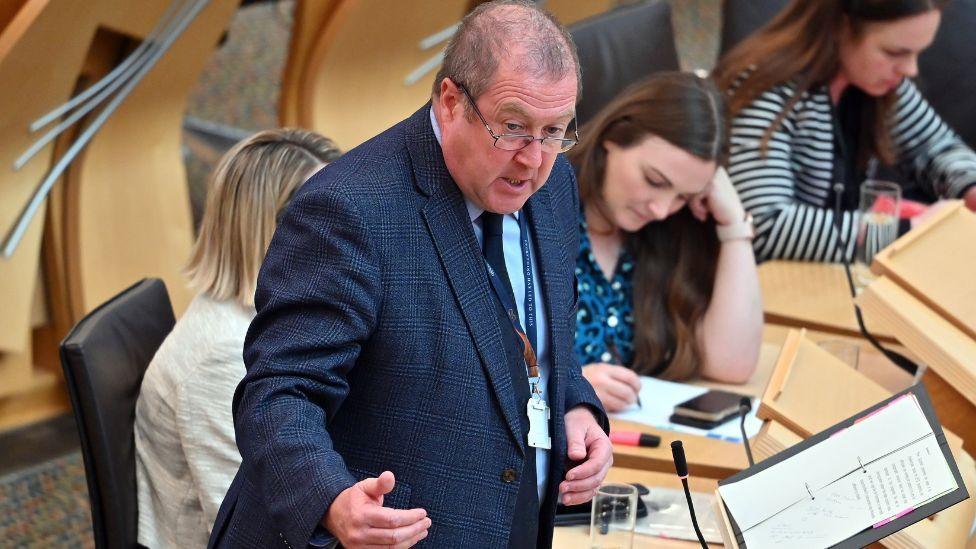 A man with thinning grey hair and glasses, wearing a blue suit, speaks in the parliament chamber. 