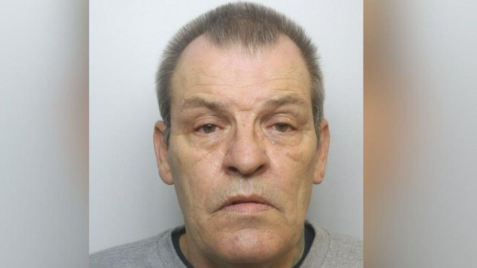 Sydney Burdon looking serious at the camera for his mug shot picture. He has a receding hairline and brown hair, wearing a grey top on.