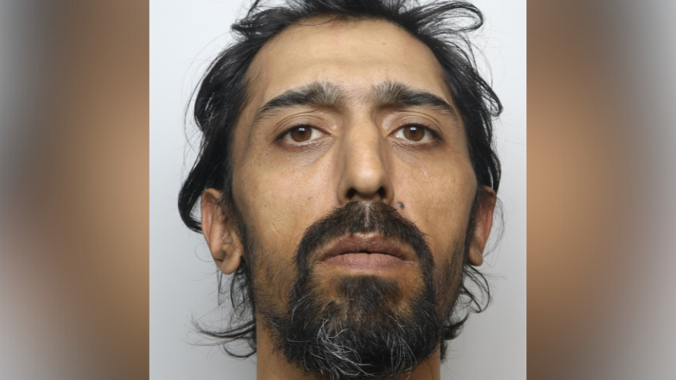 A close-up portrait of a man with dark hair and a beard, looking directly at the camera against a plain background.