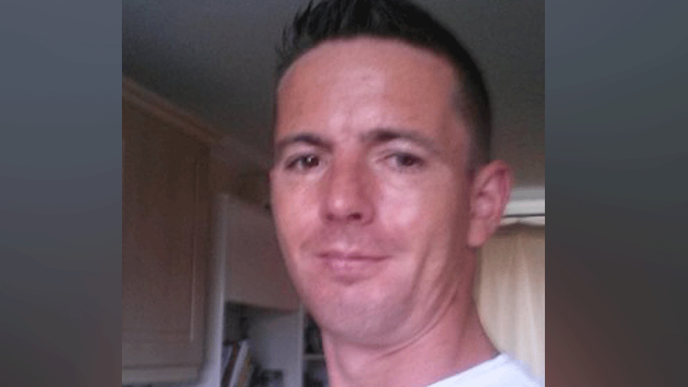 A close up selfie-style photograph of Michael Griffiths. His head is visible from the shoulders up. He has short dark brown hair and brown eyes. He appears to be stood in a room inside a house. 