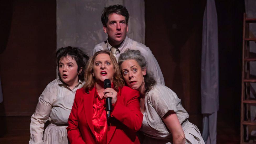 Still photography from Faust-ish play - a woman with blonde hair, in red silk pyjamas, sings into a microphone. Beside her a man and two women, both in grey bedwear, stand close to her, looking shocked. The staging is minimal, with several sheets draped across a red brick background.