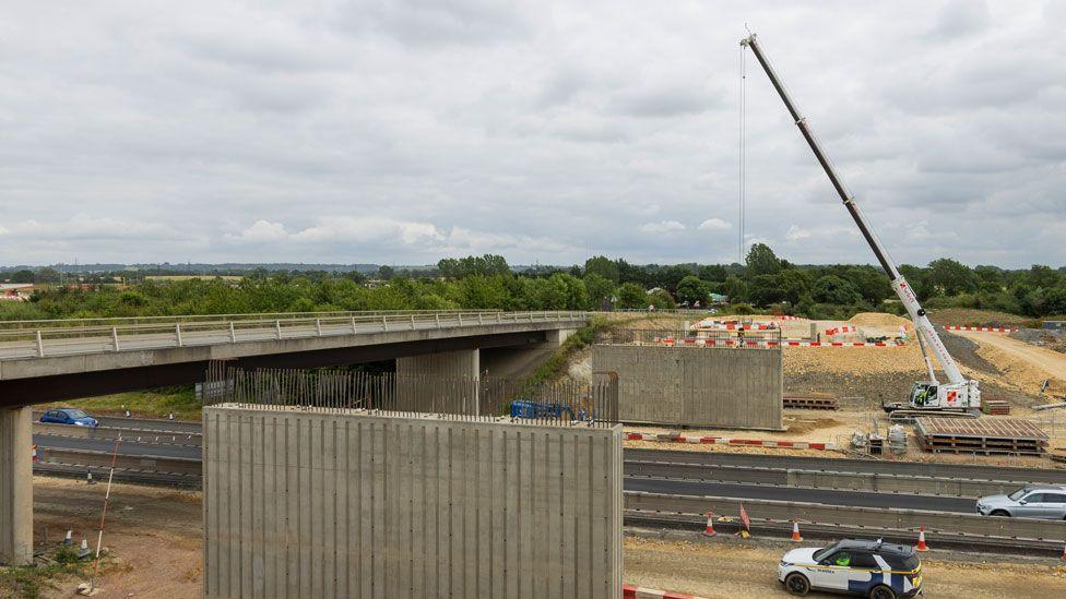 A grey concrete bridge over a dual carriageway. On either side of the dual carriageway construction is under way. The far side is busy, with a white crane and piles of road building materials. The near side is coned off but no work is being undertaken.