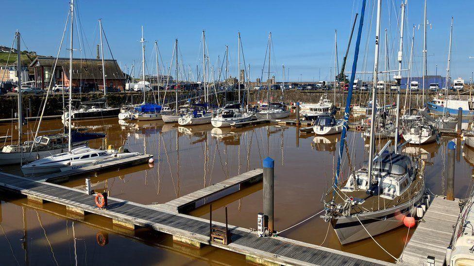 More than a dozen small boats moored at Whitehaven Harbour. The water is a dark orange/brown colour.