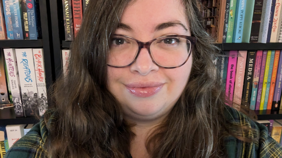 Bethan Hindmarch who has long dark hair and glasses and is wearing a green check shirt, in front of a bookcase full of books