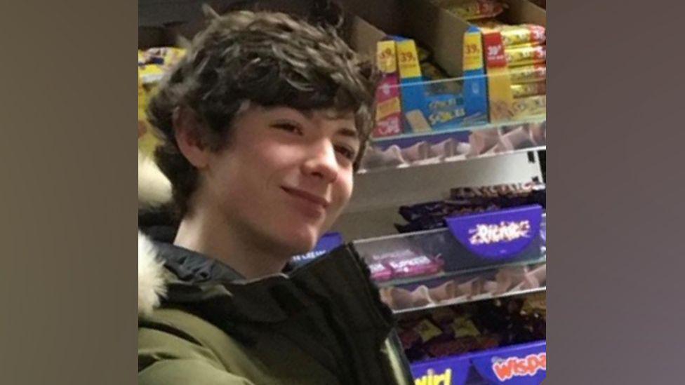 Kasey has short brown hair and is stood in a shop in front of chocolate bars. He is wearing a green coat with a white fur-lined hood