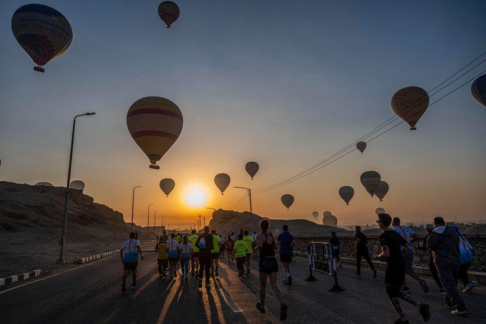 The sun rises in the sky filled with hot air balloons as people run along a road as part of the Egyptian Marathon.