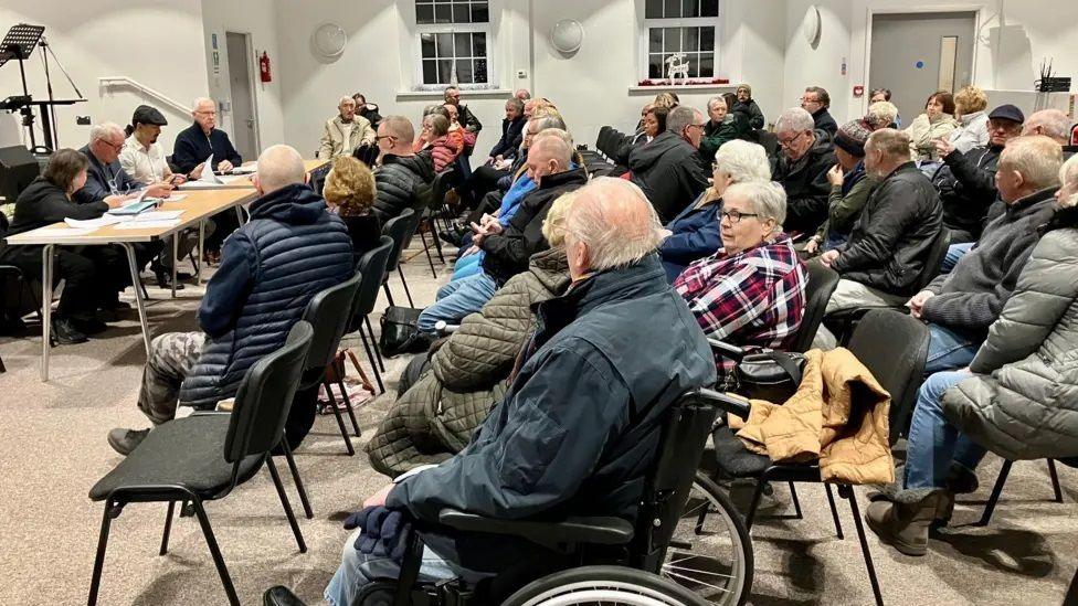 Members of the public are sat on five rows of black plastic chairs facing a row of four people sat at a table. Many of those in the hall for the public meeting are elderly.