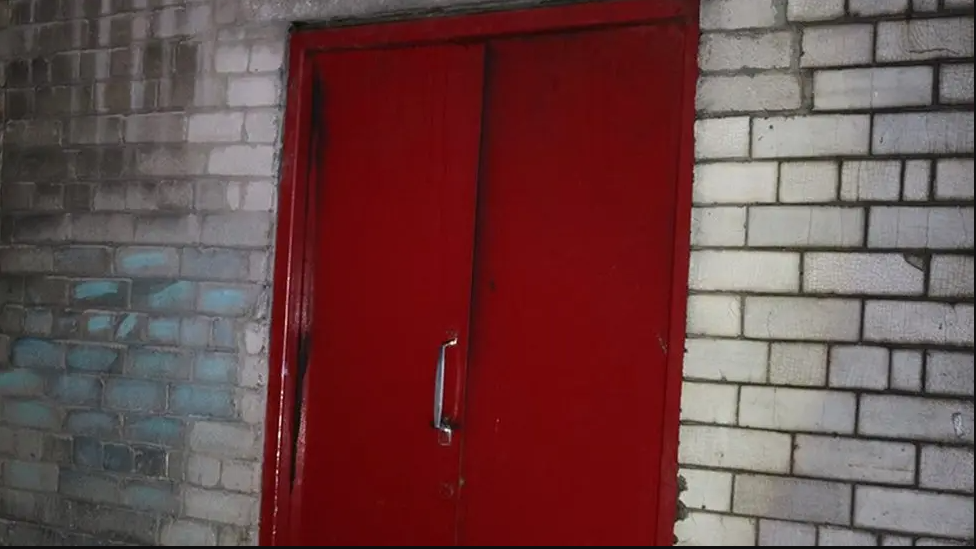 A red fire safety door in a brick wall. The fire safety door has a metal handle