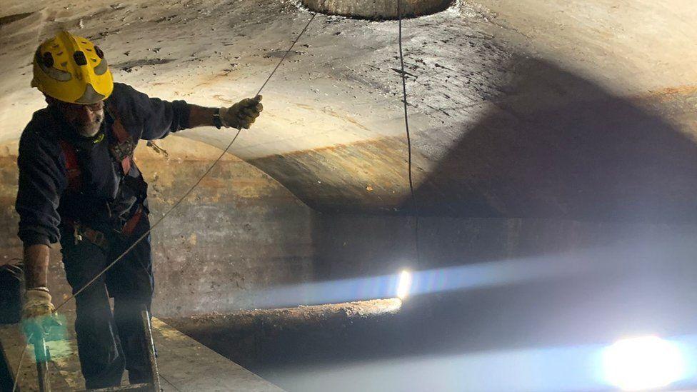 A worker in overalls and a yellow safety helmet stands on a parapet inside an underground tank which is lit by bright lamps