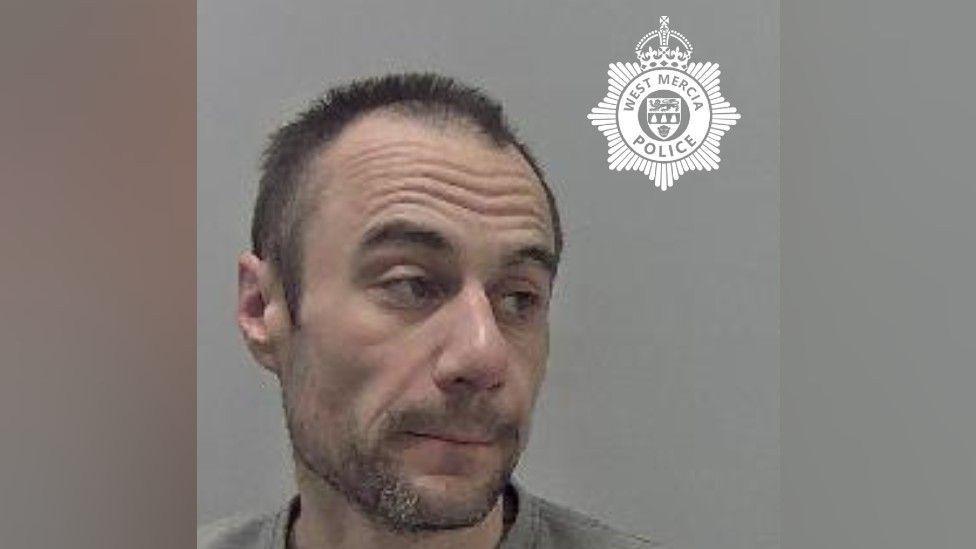 A mugshot of a man who has short black hair and dark stubble. He is wearing a grey jumper and looking to the side in a police mugshot. There is a West Mercia Police logo in the top right of the image. 