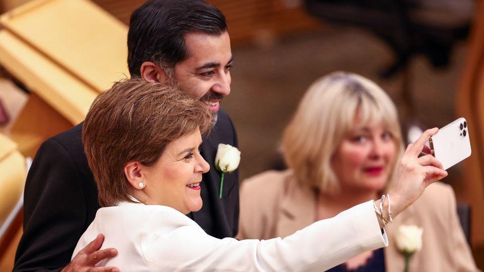 Nicola Sturgeon, with short brown hair and wearing a white suit, holds a white phone in front of her to take a selfie with Humza Yousaf, wearing a dark suit and standing next to Sturgeon with a hand on her back, in the Scottish Parliament. 