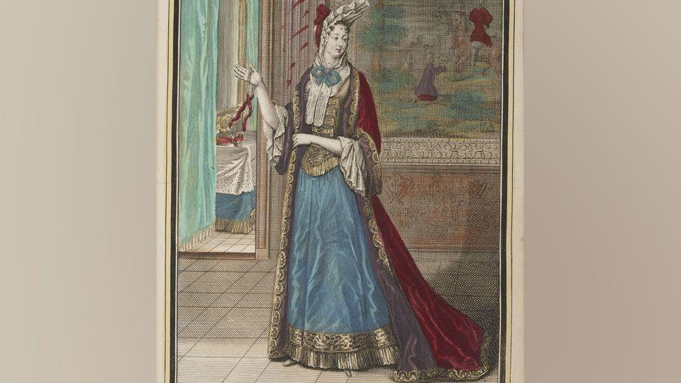 A late 17th Century woman wearing a white headdress held in place by a dark red scarf around her head and a white lace and blue ribbon around her neck and a dark red gold trimmed over dress on top of a gold bodice and sky blue gold-trimmed skirt with white lace sleeve cuffs