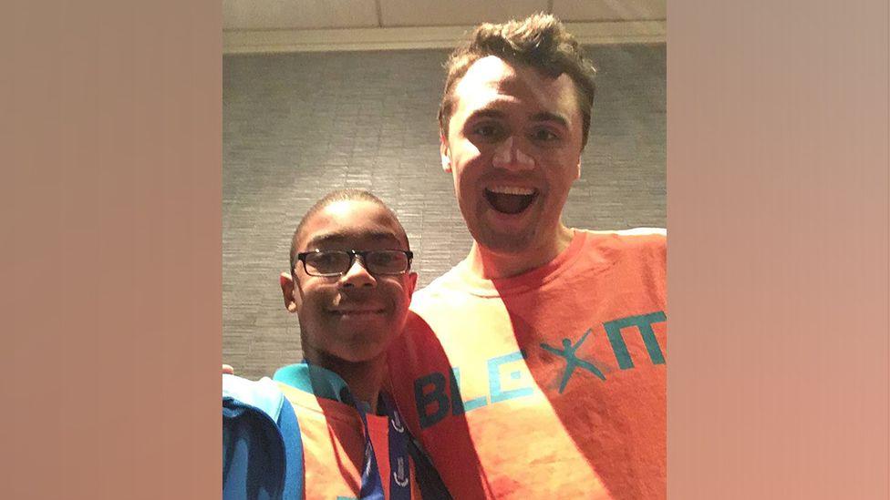 Chandler Crump, wearing glasses, a blue hoodie and a bright orange t-shirt, smiles in a selfie-style photo with Charlie Kirk, who is wearing the same orange t-shirt.