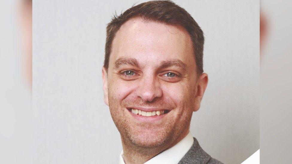 A head and shoulders shot of a man smiling into the camera. He has short brown hair and is wearing a grey blazer and white shirt.