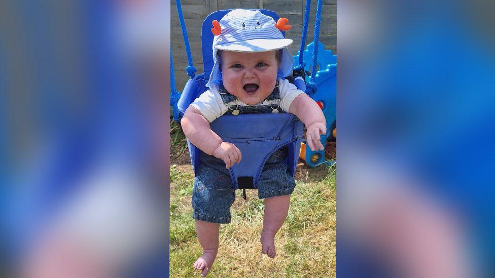 A baby of about 1 year old sitting in a baby swing in a garden in the sunshine. He is wearing a blue and white striped cap and blue denim dungarees over a white t-shirt and his feet are bare. He is looking at the camera and laughing. The grass is visible underneath him and there is a grey garden fence behind.