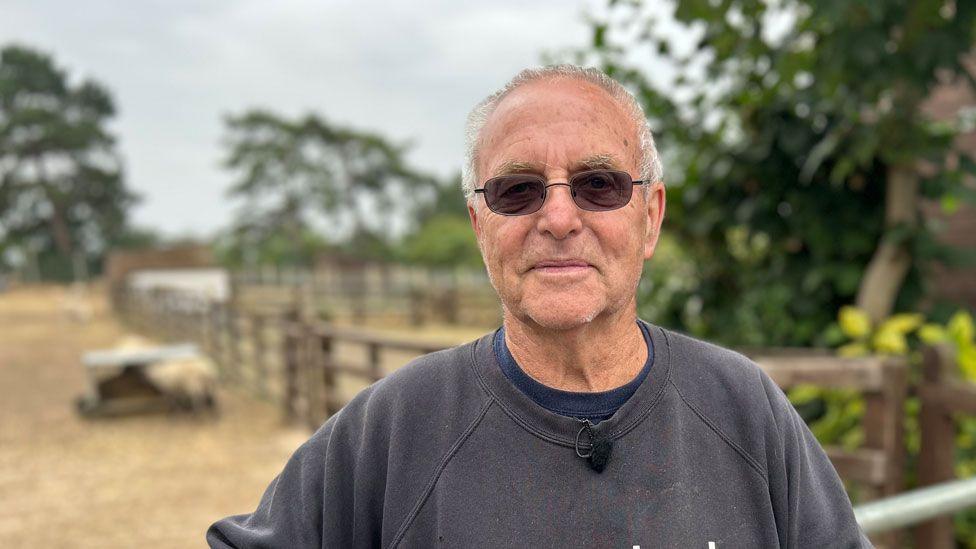 Roy Marriott with short white hair, wearing dark glasses and a grey sweatshirt over a blue t-shirt smiling at the camera and standing in front fenced off fields