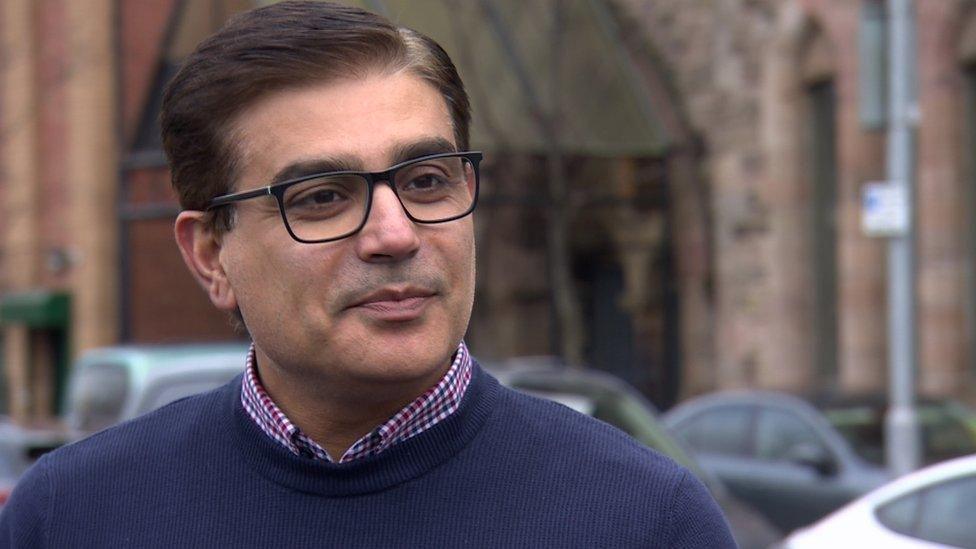 Muhammad Atif is looking off to the left. He has brown hair and is wearing black glasses, a navy jumper with the collar of a checked, purple and white shirt visible underneath. The backdrop is the Belfast Multi-Cultural Association building -a brown brick church-style build with an arched entrance. There are also a number of cars visible parked on the street.
