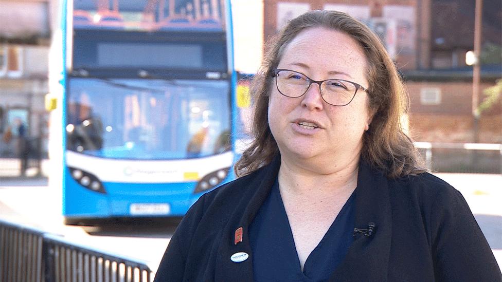 Kerri Cheek stands in front of a blue Stagecoach bus. She has brown hair, glasses, and is wearing a blue top under a black coat.