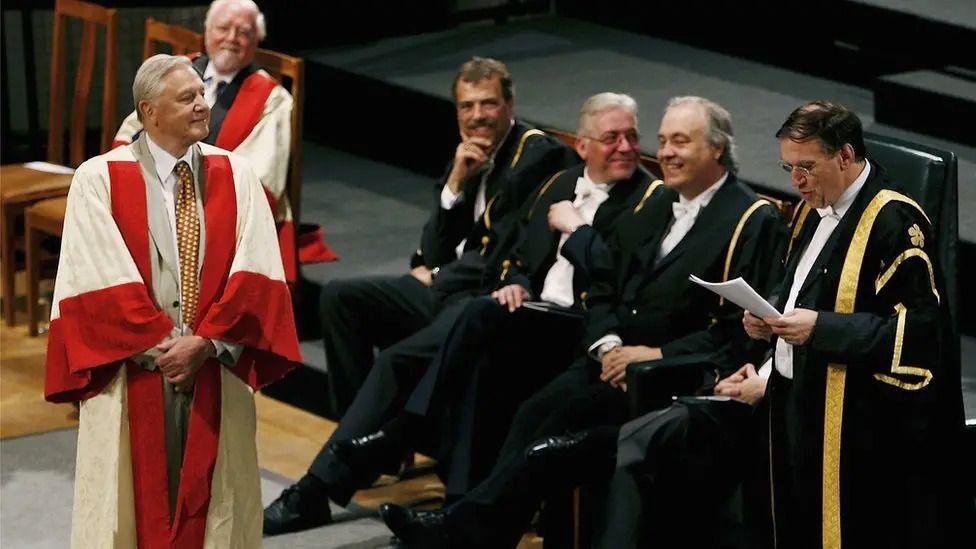 David Attenborough on the left wearing a white and red gown on stage with other university people in black gowns with a yellow trim.