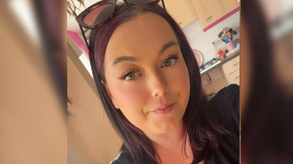 Close up of Shannon's face, she has brown hair and eyes and sunglasses on her head. She is stood in front of an open-plan kitchen.