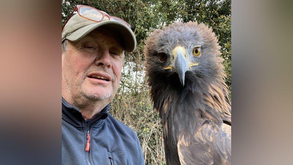 A selfie of Steve, who is wearing a cap with glasses on top of it and a navy jumper. He is next to a large bird of prey with a yellow and black beak and brown feathers.