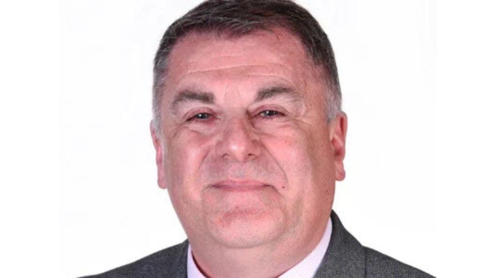 A man with dark greying hair looks at the camera in a close-up shot. He has dark eyebrows and is wearing a dark grey suit and light-coloured shirt. He visible from the neck up. He is in front of a white background.