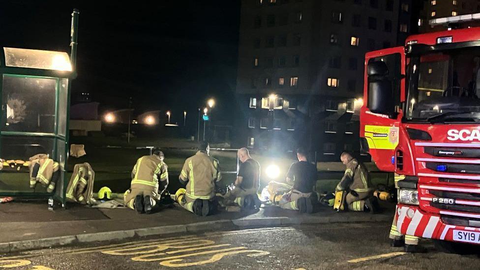 Several firefighters in uniform, in the dark, next to a red fire engine with its door open.