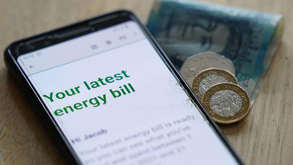 Generic photo of a mobile phone showing an online energy bill on a table next to a five pound note, two pound coins and a 50p piece