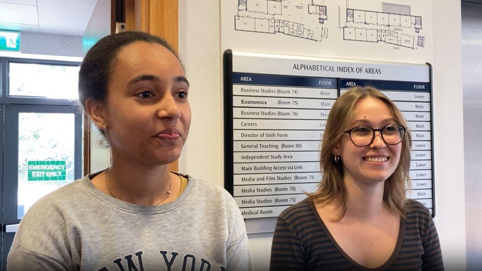 Gracia van Zutphen and Lea Ackermann stand in a college hallway. Gracia has black hair, which she is wearing up, is has on a light grey sweatshirt. Lea has shoulder-length fair hair is wearing glasses, and is in a dark striped top.