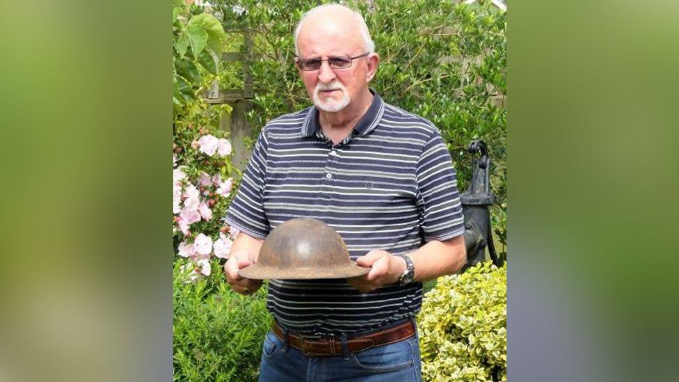 Dennis O'Callaghan, with white hair, beard and glasses, wearing a blue striped short-sleeved shirt while stood in a garden. He is holding a World War One British helmet, which was refurbished in the late 1930s and used during World War Two.