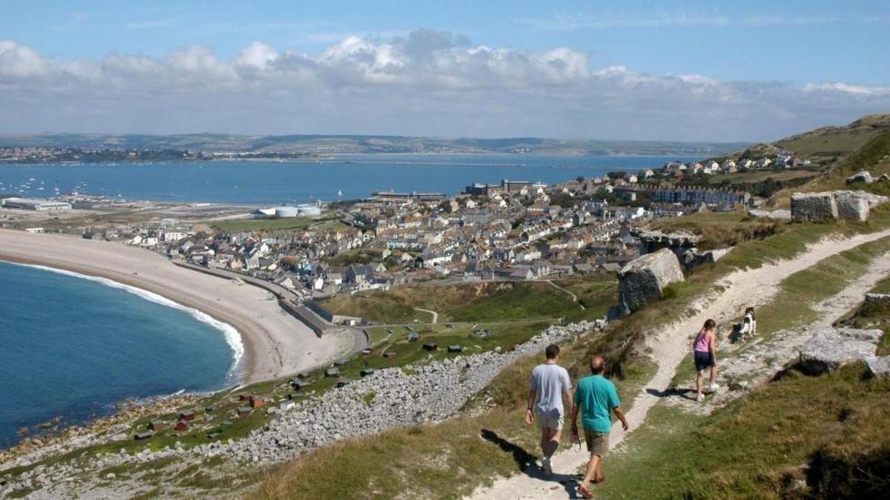 Elevated view from the top of Portland across Portland Harbour, Chesil Beach and Weymouth in the distance on a bright sunny day. In the foreground is a group of people walking on a with a dog.