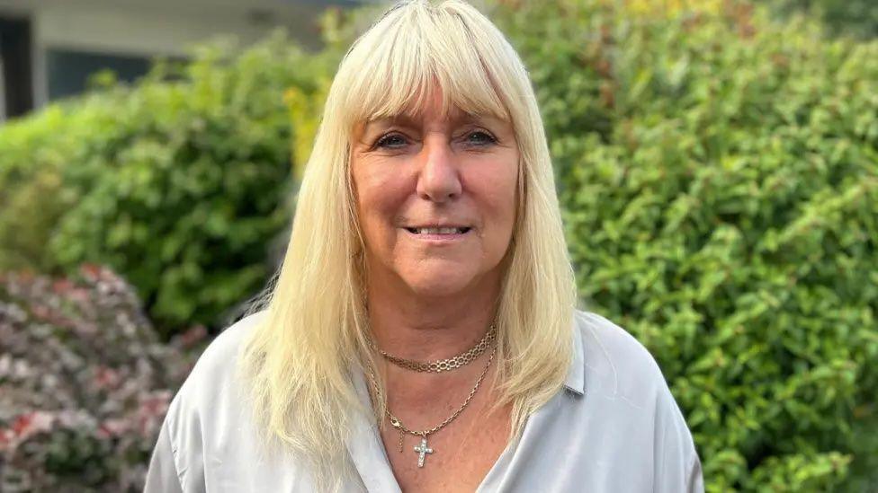 Julie, a woman with blonde hair, she has a fringe and wears a white top with some necklaces as she stands in front of a green bush.