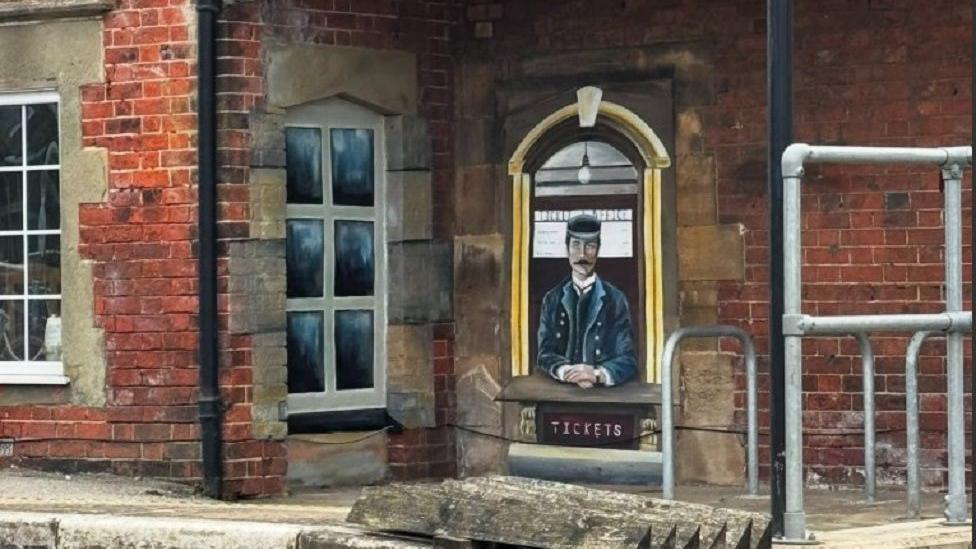 How Nikita Spires' mural looked when she finished it, viewed on the wall of the station from a distance. On the left hand side, she has painted a window that looks like the others, complete with grey stones around it and six large panes. On the right hand side, she has painted an old-fashioned, Victorian ticket officer wearing a navy uniform and flat cap, sat waiting for tickets.