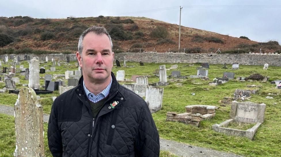 Adrian Hughes stands in a cemetery with headstones in the background