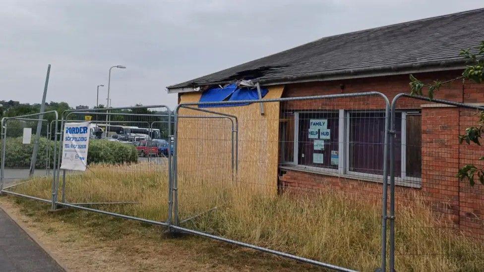 A building has wood boarded over a part of it, and has a partially collapsed roof. There is metal fencing around the building and blue tarpaulin over a gap in the wall