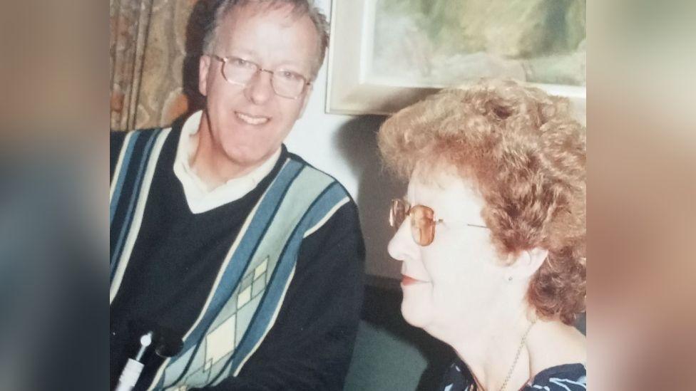 Michael "Mick" Page and Elizabeth Page pictured together seated indoors. Mick Page is on the left is wearing glasses, a dark-coloured sweater with an argyle pattern, and is smiling. Elizabeth Page is on the right, wearing glasses and has curly hair. She is dressed in a dark, floral-patterned blouse.