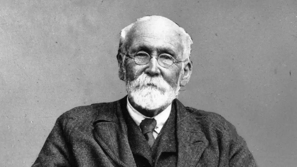A black and white image of an old man wearing a suit. He has a white beard and moustache and is wearing small circular glasses.